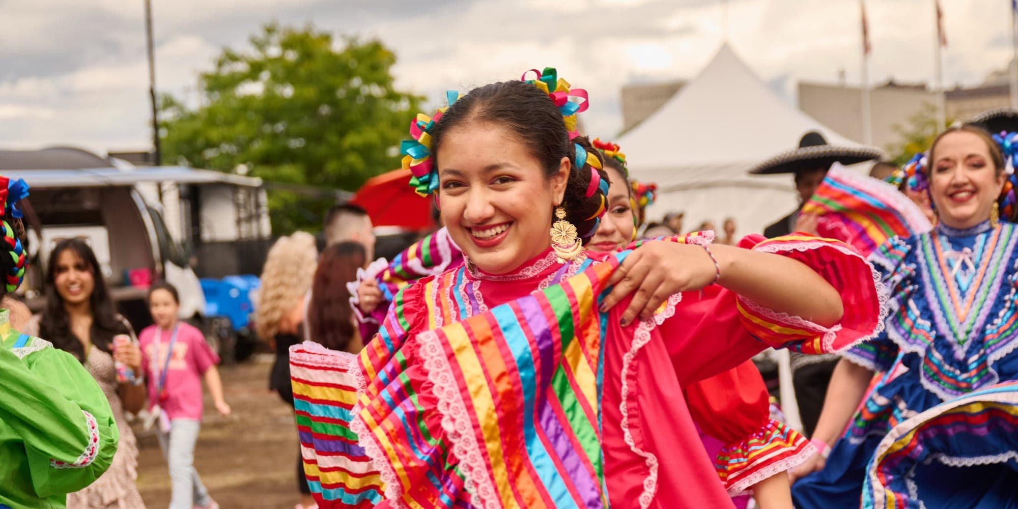 A folklórico dancer in a pink dress striped with rainbow ribbons swirls her skirt and smiles, with other dancers behind her.