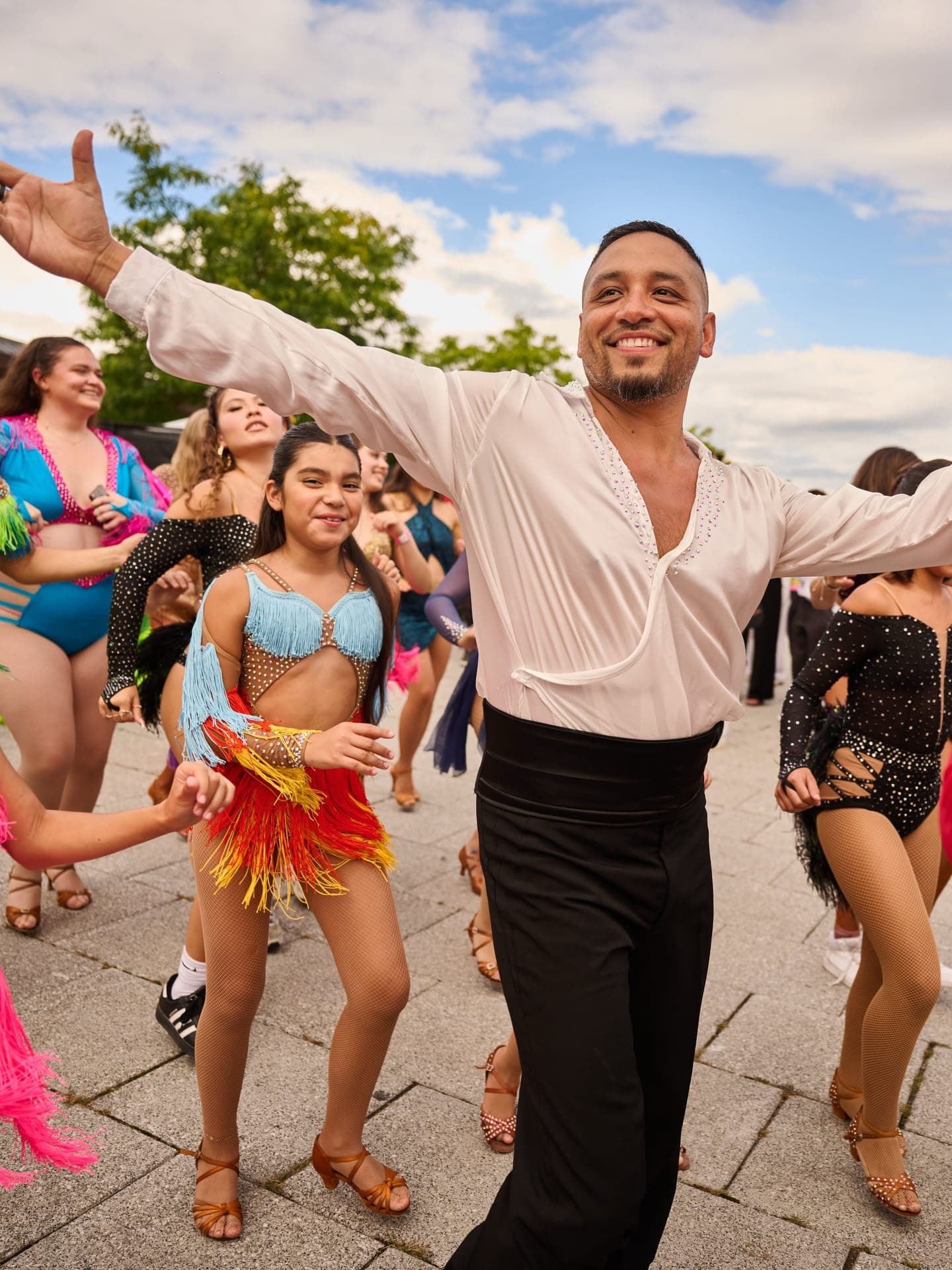 A dancer in a white ballroom shirt spreads his arms wide, smiling, leading a line of costumed dancers across the plaza at Latin Sparks in Ottawa.
