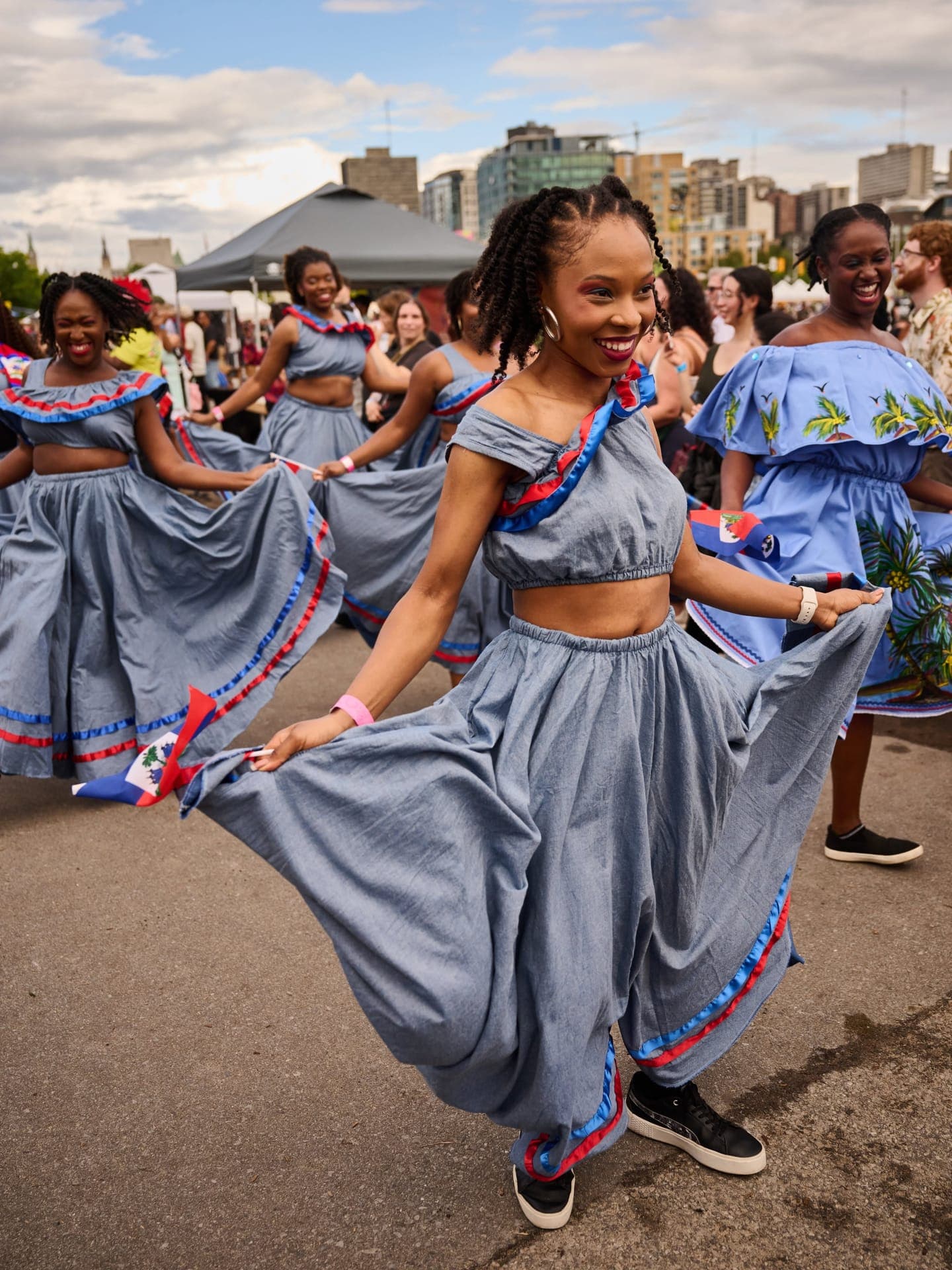 Dancers in denim-blue dresses trimmed with red and blue ribbon twirl their skirts and wave small Haitian flags, with the Ottawa skyline behind them.